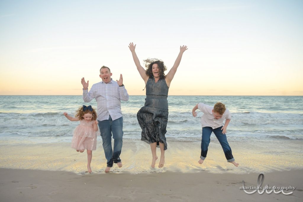 Four people jump along a sandy shoreline at sunset, with ocean waves, wet sand reflections, open sky, and a beach photography watermark visible in the lower corner