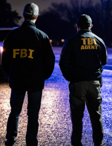 Two officers at night with backs to camera; one in 'FBI' jacket, the other in 'TBI AGENT' jacket, standing in dark outdoor setting with blurred background