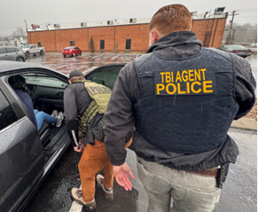 TBI agent in navy jacket assists tactical officer helping person from black car in rainy parking lot; brick building and vehicles in background.