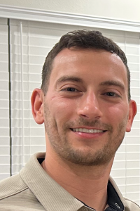 Person in beige Under Armor polo stands before white blinds, with white wall and ceiling in background