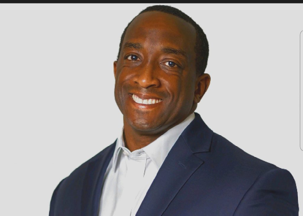 Individual in navy blue suit jacket and white dress shirt posed against plain background