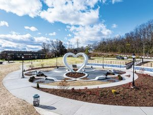 Circular memorial plaza with curved concrete walkways, landscaped flower beds, benches, and a large white heart-shaped sculpture at center, set within a park with trees, nearby roads, and open sky.