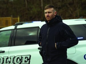 Officer wearing a black weather‑resistant jacket stands beside a marked police SUV with roof lights, duty belt and radio visible, with wooded background