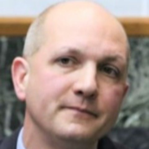Close‑up portrait in indoor setting shows formal attire including dark suit jacket, collared shirt, and tie against a softly lit background wall panel