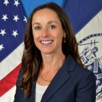 Individual wearing a navy blazer over a white textured top, gold necklace, and drop earrings stands before U.S. and agency flags, with long dark hair, badge-style seal, blue backdrop, and professional portrait lighting visible throughout the formal composition