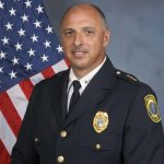 Police chief in formal dark uniform with gold buttons, badge, and shoulder insignia, standing before a U.S. flag against a gray studio backdrop