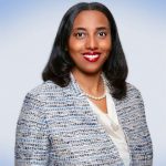 Individual in blue and white textured blazer, white blouse, and pearl necklace stands before a smooth gradient blue background