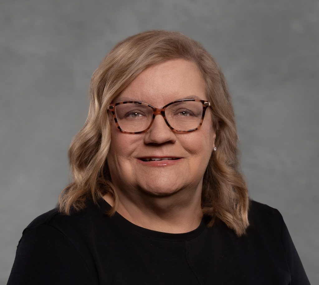 Person with shoulder-length wavy blonde hair wearing a plain black top, posed against a smooth gray studio background with soft lighting