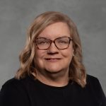 Person with shoulder-length wavy blonde hair wearing a plain black top, posed against a smooth gray studio background with soft lighting