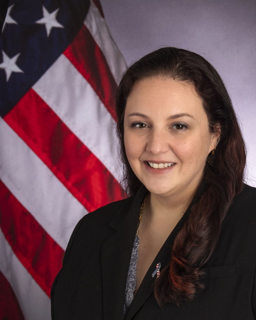 Studio portrait of a professional wearing a dark blazer over a patterned blouse, posed before a gray backdrop with a vertically displayed U.S. flag to one side