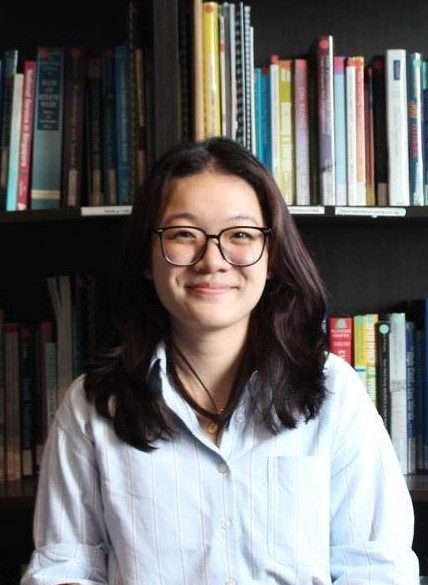 Individual wearing a dark blazer over a dark top sits before bookshelves filled with books and binders, with library shelving, colorful spines, and reference materials visible throughout the background
