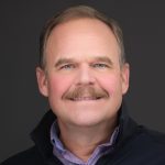Person in dark jacket and light purple checkered shirt, collar visible, posed against a solid dark background