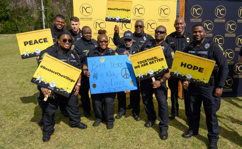 “Group of uniformed police officers standing outdoors holding yellow signs reading ‘Peace,’ ‘Hope,’ and ‘Together We Are Better,’ with banners behind them.”