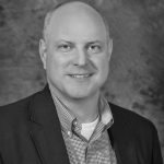 Black and white image of person in suit jacket and checkered shirt posed against textured backdrop, emphasizing professional attire and monochrome tone