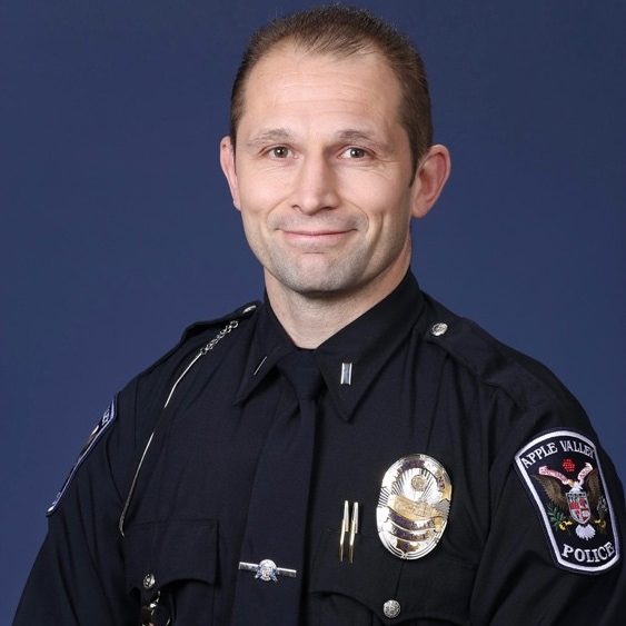 Apple Valley Police officer in uniform with visible badges and insignia stands against blue backdrop.