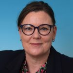 Person wearing a dark blazer over a patterned shirt with pink and green shapes, set against a solid blue background in a professional portrait style.