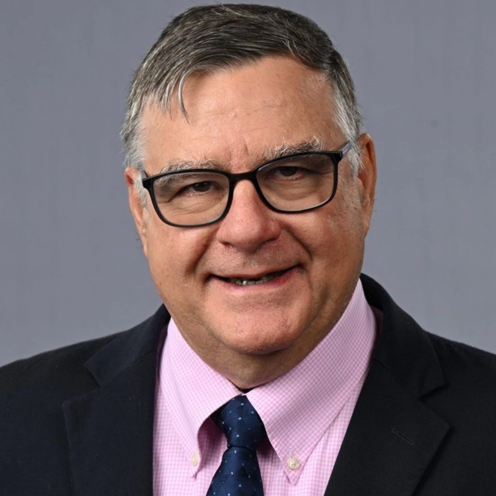 individual with short, dark hair and glasses. They are wearing a dark suit jacket over a light pink dress shirt. The individual has a navy blue tie with small white polka dots. The background is plain gray.