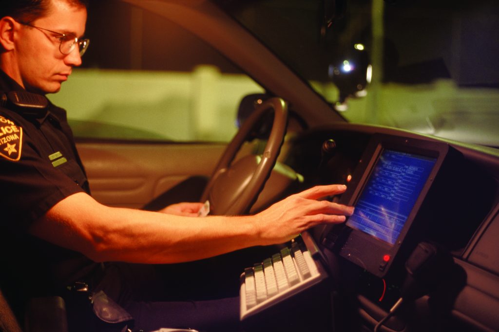 Police officer inside patrol car using mounted touchscreen computer with visible keyboard, steering wheel, and dashboard controls in dim lighting