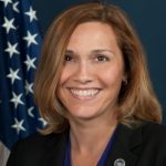 Person in dark blazer with blue blouse and lapel pin stands before a U.S. flag on a dark blue background, suggesting an official or professional setting