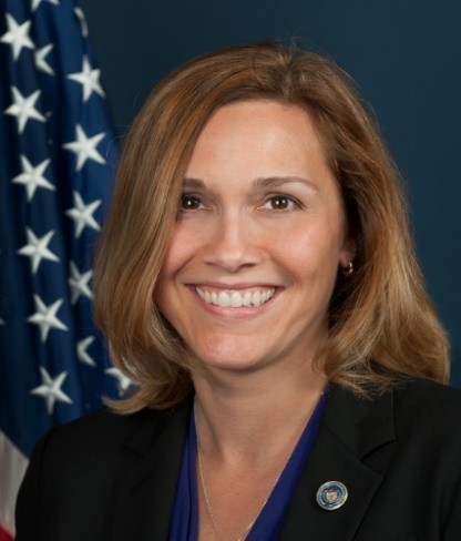 Person in dark blazer with blue blouse and lapel pin stands before a U.S. flag on a dark blue background, suggesting an official or professional setting