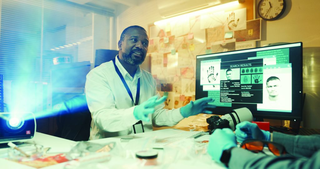 Forensic investigator in lab coat and blue gloves analyzing evidence at desk with camera, documents, and computer displaying fingerprint search results.