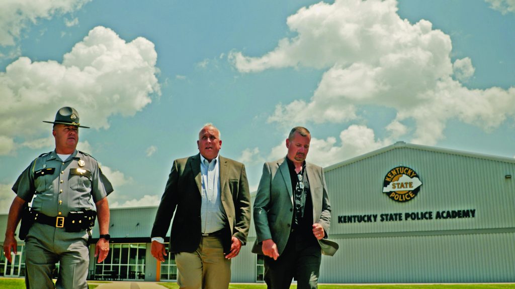 Three individuals in formal and police uniforms walk outside Kentucky State Police Academy building under a bright sky with scattered clouds