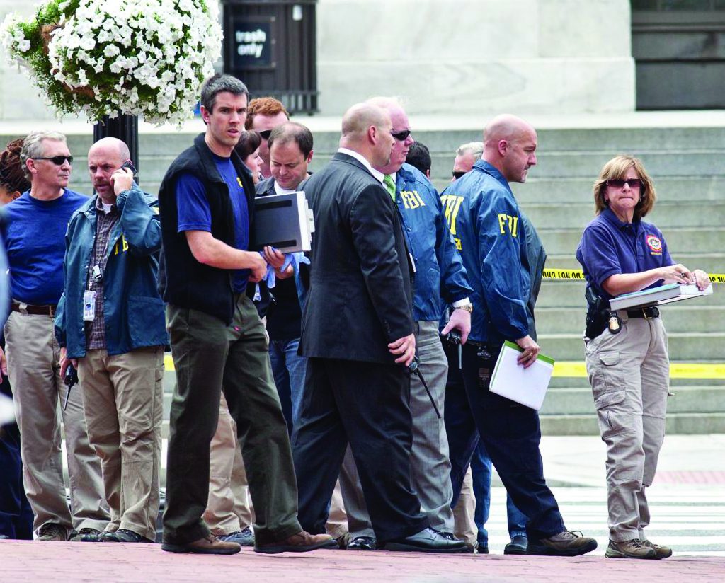 A group of personnel in tactical and professional attire stand near steps and caution tape, reviewing notes and equipment during an outdoor investigative scene