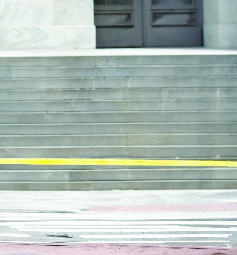 Wide concrete steps lead to large double doors on a stone building, with yellow caution tape stretched across the lower steps above a crosswalk and paved street