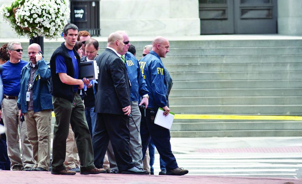 A group of investigators and personnel stand on a street near concrete steps marked with yellow tape, holding documents and equipment as they gather beside parked vehicles