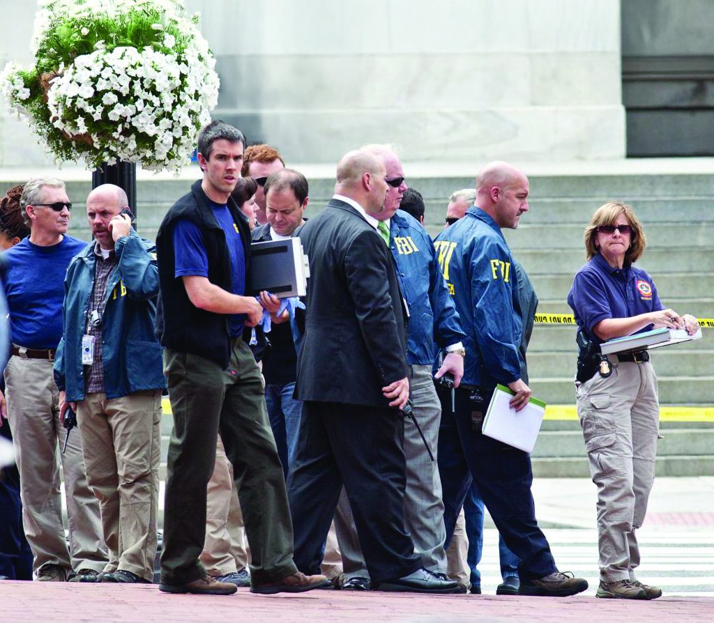 A group of investigators and uniformed personnel stand near concrete steps marked with yellow tape, holding documents and equipment beside a hanging planter.