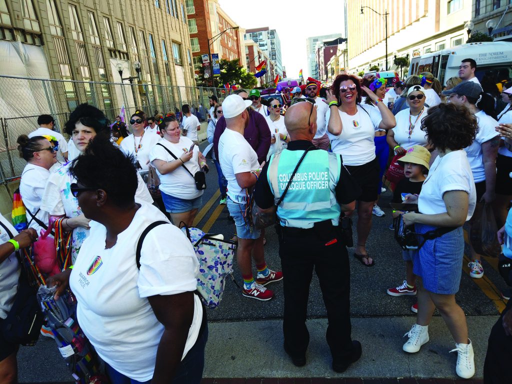 Crowd gathers at a city street parade with rainbow flags, a Columbus Police dialogue officer in a blue vest, participants in white shirts, vehicles, and buildings lining the route