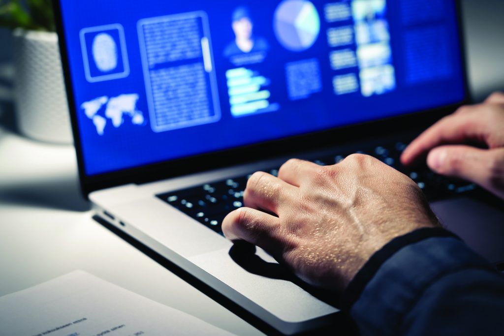 Close-up of hands typing on a laptop with dark screen displaying fingerprint graphic, profile data, charts, and world map in a dimly lit workspace