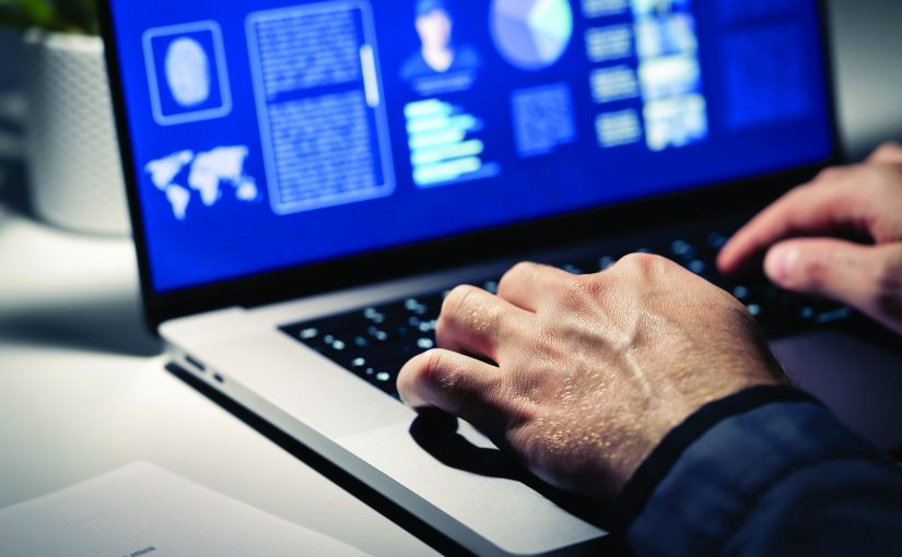 Close-up of hands typing on a laptop with dark screen displaying fingerprint graphic, profile data, charts, and world map in a dimly lit workspace