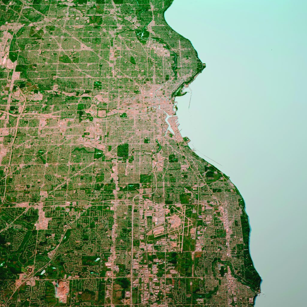 Aerial view of an urban grid layout along a curved shoreline with dense streets, green patches, and Lake Michigan’s light blue water on the right edge