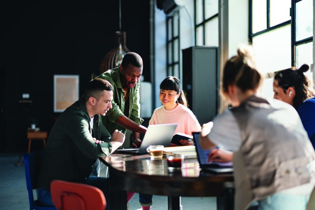 Group of people gathered around a wooden table in a modern office with large windows, laptops, notebooks, and coffee cups during a collaborative meeting