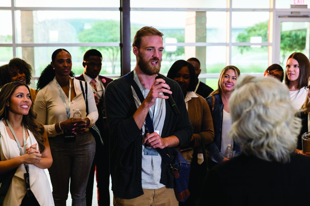 Group of people standing indoors near large glass windows, one person holding a microphone and speaking, others wearing lanyards and holding drinks