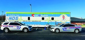 Two police vehicles parked in front of large NDS-branded 'Heart Mobile' health unit with green-blue design and contact info; mobile care support visible