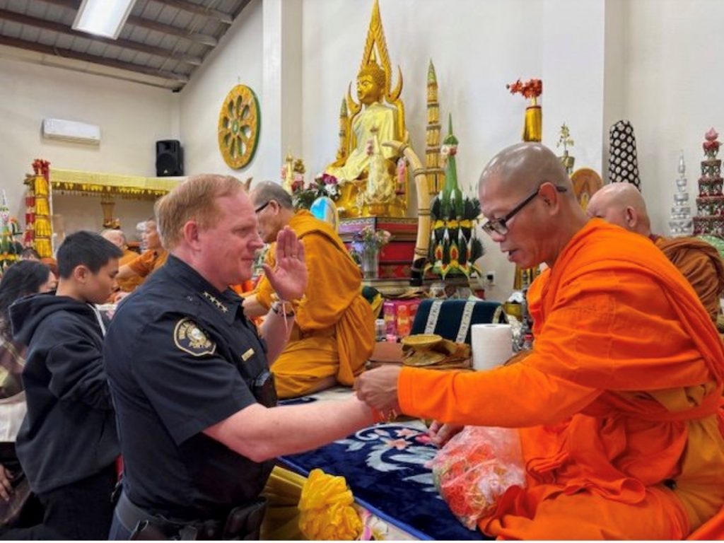 Portland Police officer in dark uniform participates in a cultural ceremony inside a temple, exchanging items with monks in orange robes seated before ornate golden statues and decorative offerings.