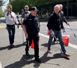 Uniformed Portland Police officer and community members walk on a city street carrying red buckets and litter pickers, wearing gloves, participating in a cleanup event.