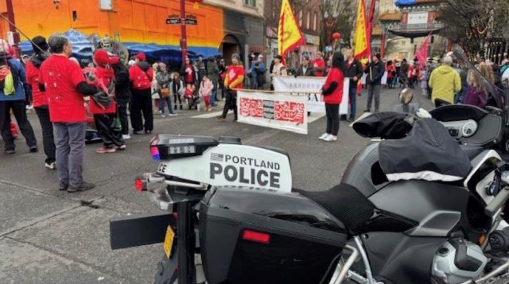 Portland Police motorcycle parked near a street parade with participants in red shirts holding banners and yellow flags, colorful mural and Chinatown gate in background.