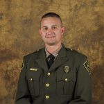Uniformed officer in green jacket with gold buttons, badge, and shoulder patches sits against a brown mottled backdrop; formal portrait-style setting